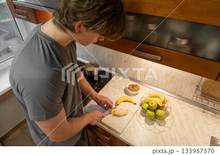 Cutting fruit caucasian man in home kitchen interior preparing healthy breakfast, indoor lifestyle scene with fresh food and nutrition. Morning Kitchen Routine: Cooking Oatmeal and Coffee Cutting fruit caucasian man in home kitchen interior preparing healthy breakfast, indoor lifestyle scene with fresh food and nutrition. Morning Kitchen Routine: Cooking Oatmeal and Coffee 133987370