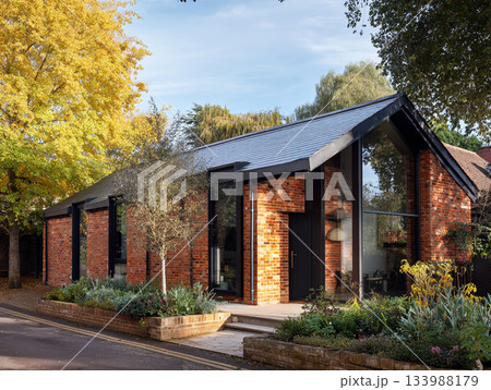 A modern luxury house featuring dark red brick, black-framed windows, a slate roof, and a large integrated garage, surrounded by lush mature trees and a paved driveway. 133988179