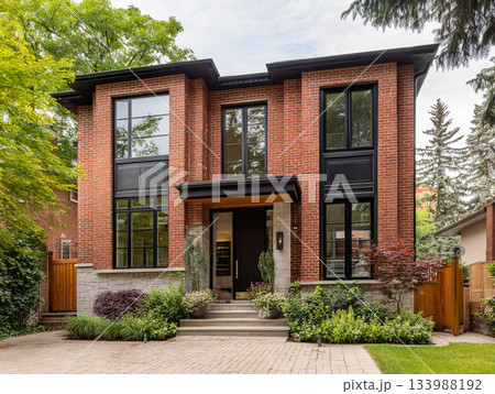A modern luxury house featuring dark red brick, black-framed windows, a slate roof, and a large integrated garage, surrounded by lush mature trees and a paved driveway. 133988192