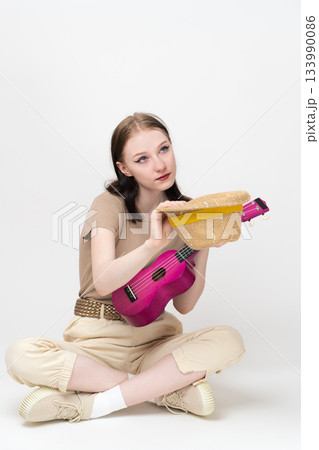 Female musician with ukulele holds straw hat in front of her for donations and tips, sitting cross legged on floor, looking up. Woman ukulele artist Caucasian ethnicity in casual, comfortable outfit Female musician with ukulele holds straw hat in front of her for donations and tips, sitting cross legged on floor, looking up. Woman ukulele artist Caucasian ethnicity in casual, comfortable outfit 133990086