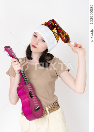 Young female musician attired in Santa Claus hat and she holds ukulele in one hand and pom pom in other. Her facial expression is characterized by playful and friendly demeanor, as she looks at camera 133990089
