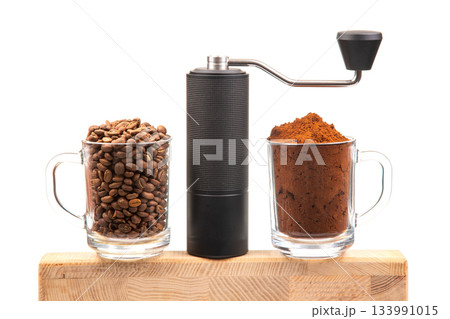 A manual coffee grinder sits between two glass mugs, one with roasted coffee beans 133991015