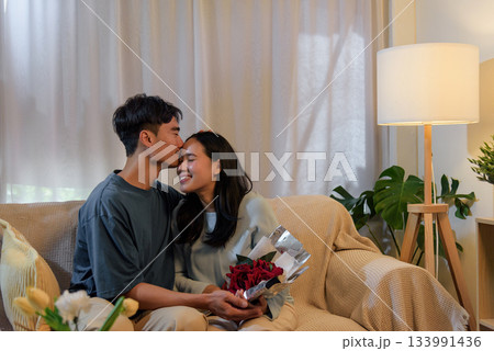 Young couple happy love, man woman share romantic valentine day at home, man kisses woman on forehead while she holds beautiful red roses, perfect moment expressing affection. 133991436
