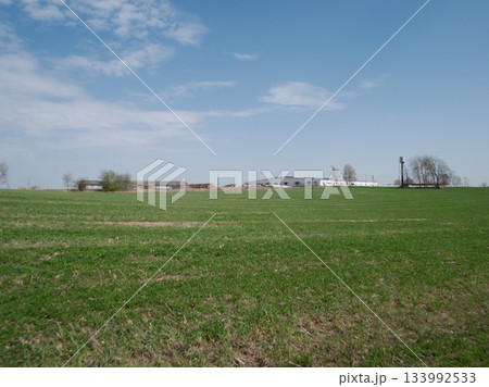Rolling green grasses stretch across the countryside, framed by agricultural structures and a clear blue sky, depicting a serene rural setting. Rolling green grasses stretch across the countryside, framed by agricultural structures and a clear blue sky, depicting a serene rural setting. 133992533