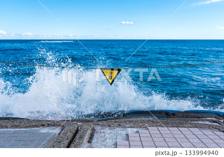 海辺に掲げられた注意標識と波しぶき 133994940