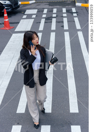 Young Asian woman doing business work in suit on street, busy professional talking on phone, holding laptop and travel mug, navigating urban environment, focused on important tasks. Young Asian woman doing business work in suit on street, busy professional talking on phone, holding laptop and travel mug, navigating urban environment, focused on important tasks. 133999859