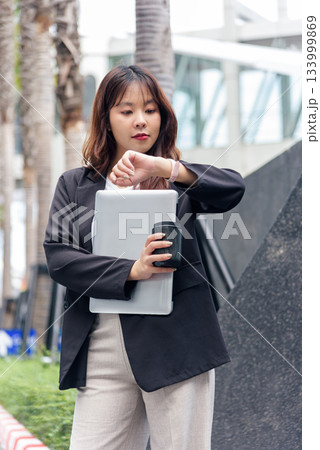 Young Asian business woman, busy checking watch on street, professional work suit, holding modern laptop, coffee cup, urban cityscape background, confident office employee, ready for meetings. 133999869