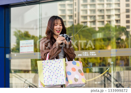 Asian woman enjoys shopping, happy buyer holds stylish fashion bags, posing with camera outside modern city mall, capturing exciting black friday lifestyle, people love great deals. 133999872