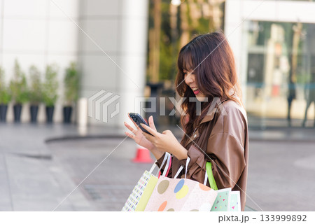 Asian woman enjoys shopping, active lifestyle for people, holding fashion bags near mall entrance, smiling brightly, posing outdoors after black friday shop event, checking mobile phone. Asian woman enjoys shopping, active lifestyle for people, holding fashion bags near mall entrance, smiling brightly, posing outdoors after black friday shop event, checking mobile phone. 133999892