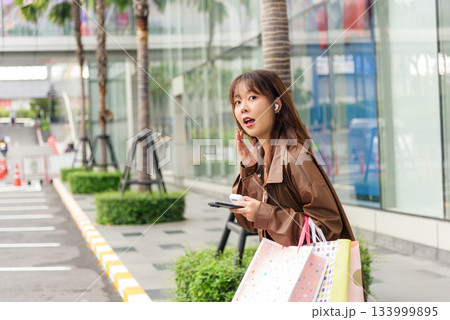 Asian woman enjoys shopping lifestyle, holding fashion bags and black phone in hand posing near modern city mall, people notice stylish urban look showing joy from purchases. Asian woman enjoys shopping lifestyle, holding fashion bags and black phone in hand posing near modern city mall, people notice stylish urban look showing joy from purchases. 133999895