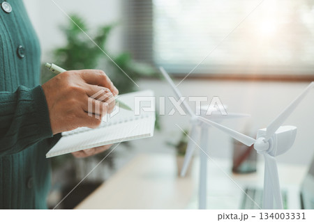 Eco-friendly office scene with person working on laptop near solar panel and wind turbine model, representing green business, renewable energy innovation and sustainable technology workspace. 134003331