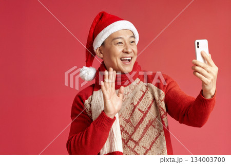 Man Wearing a Santa Hat Takes a Joyful Selfie During the Holiday Season Against a Red Backdrop in a Festive Setting 134003700