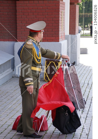 The photograph depicts a man in military uniform standing on the street, holding a red flag with a star and a hammer and sickle. Next to him is a black suitcase. The man is wearing a green uniform wit The photograph depicts a man in military uniform standing on the street, holding a red flag with a star and a hammer and sickle. Next to him is a black suitcase. The man is wearing a green uniform wit 134006826
