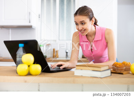 Emotional woman using laptop for work or study in home kitchen 134007203
