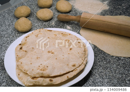 Making the unleavened flatbread roti. Close-up of several cooked roti on a white plate. Nearby are flour, a rolling pin, and balls of dough on a gray granite kitchen surface. Backing homemade bread. 134009486