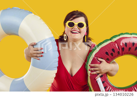 Smiling fat woman in swimsuit, sunglasses and earrings holding two inflatable beach rings 134010226