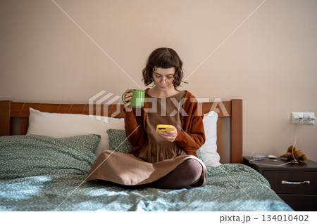 Pensive teenager sitting on bed with cup of tea, reading news in internet, hanging in social network Pensive teenager sitting on bed with cup of tea, reading news in internet, hanging in social network 134010402