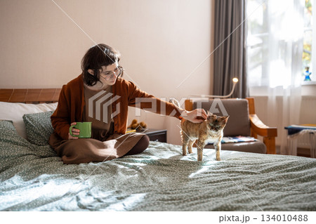 Teen girl petting Devon Rex cat sitting in bed at home drinking morning coffee. Pet lover Teen girl petting Devon Rex cat sitting in bed at home drinking morning coffee. Pet lover 134010488