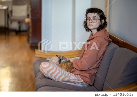 Tired pensive teenage girl petting pet cat taking break looking side sitting on couch at home.  134010518