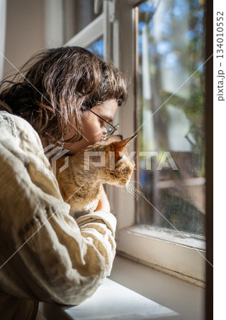 Caring pet lover cuddling beloved breed cat Devon Rex sitting on windowsill looking at window 134010552