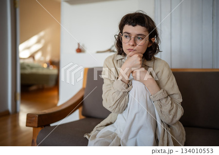 Thoughtful bored lonely teen girl in glasses feeling apathy sadly pondering sitting on couch at home Thoughtful bored lonely teen girl in glasses feeling apathy sadly pondering sitting on couch at home 134010553