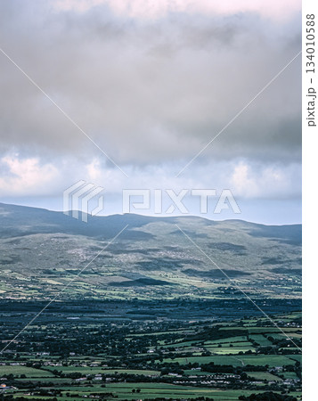 A distant view shows a valley with fields and towns below a mountain range. Clouds fill the sky above the landscape near the Coomloughra Horseshoe hiking trail. A distant view shows a valley with fields and towns below a mountain range. Clouds fill the sky above the landscape near the Coomloughra Horseshoe hiking trail. 134010588