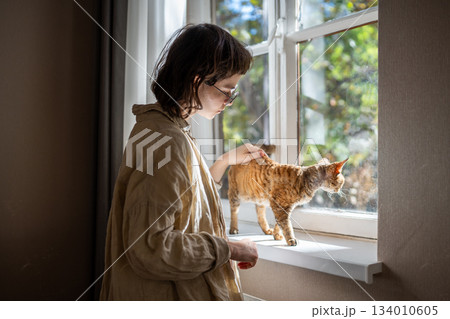 Caring lonely pet owner stroking petting curious cat on windowsill at home, enjoying interaction. 134010605