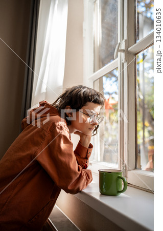 Nerd teen girl in glasses drinking tea coffee looking at window boring at home. Friendless, laziness Nerd teen girl in glasses drinking tea coffee looking at window boring at home. Friendless, laziness 134010635
