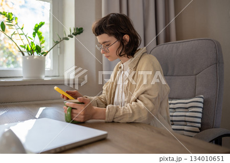 Focused girl freelancer browsing social networks looking in smartphone screen holding cup at home. 134010651