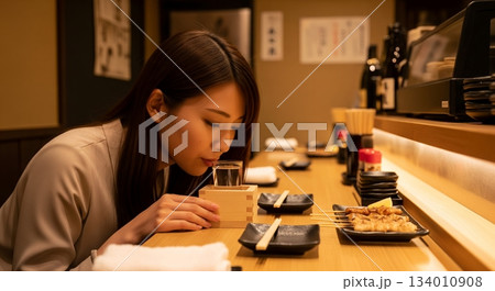 カウンター上のグラスに直接口を近づけて日本酒を飲む若い女性 134010908