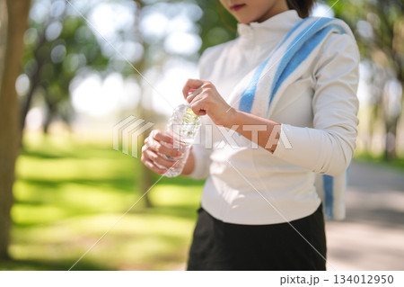 Active Asian adult woman opens water bottle refreshing body after outdoor workout in sunny park promoting hydration and healthy living practices 134012950