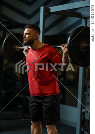man is performing squats with a barbell on his shoulders inside a gym. He focuses on his form while lifting weights. The gym is equipped with various fitness equipment. 134015072
