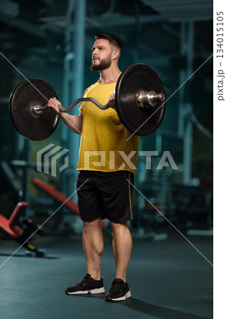 A man stands in a gym holding a barbell with both hands. He is focused on performing a bicep curl while dressed in a yellow shirt and shorts. The gym is well lit. A man stands in a gym holding a barbell with both hands. He is focused on performing a bicep curl while dressed in a yellow shirt and shorts. The gym is well lit. 134015105