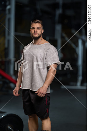 man stands focused and ready to lift a barbell for a bicep curl. He is in a gym filled with equipment. It is evening and he is dressed in workout clothes. man stands focused and ready to lift a barbell for a bicep curl. He is in a gym filled with equipment. It is evening and he is dressed in workout clothes. 134015106