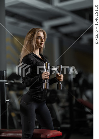 A girl lifts dumbbells while performing bicep curls in a gym setting. She focuses on her form and technique while exercising. The environment is filled with gym equipment. 134015110