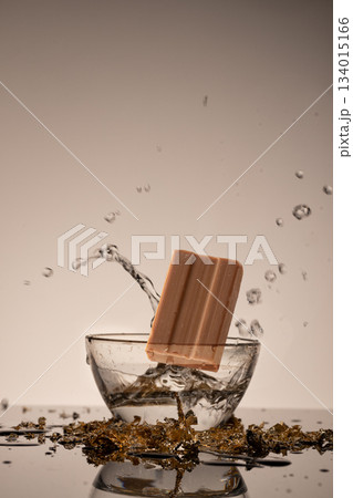block of soap drops into a glass bowl filled with water creating splashes and water droplets. The scene is set in a studio with a plain background. 134015166