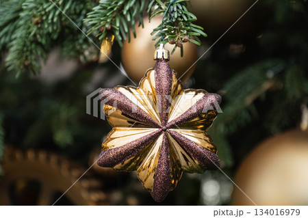 Elegant gold flower shaped Christmas ornament on green fir branches with soft holiday lights. Festive luxury decoration detail. 134016979