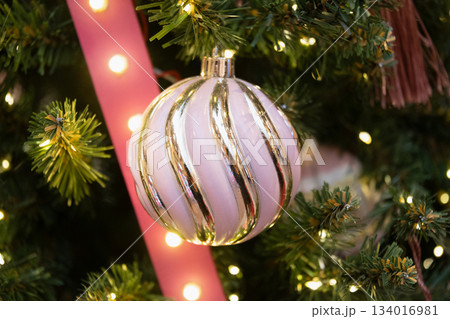 Pink and gold Christmas bauble hanging on fir tree with warm fairy lights and festive ribbon. Elegant holiday decoration. Pink and gold Christmas bauble hanging on fir tree with warm fairy lights and festive ribbon. Elegant holiday decoration. 134016981