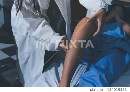 Doctor examining and supporting injured ankle of female patient in hospital room, providing medical assistance and treatment for sprain or fracture 134019331