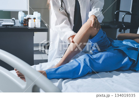 Doctor examining and supporting injured ankle of female patient in hospital room, providing medical assistance and treatment for sprain or fracture 134019335