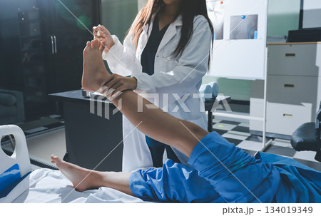 Doctor examining and supporting injured ankle of female patient in hospital room, providing medical assistance and treatment for sprain or fracture 134019349