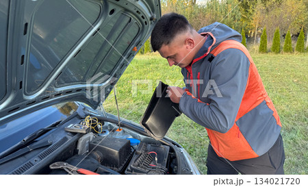 A Caucasian male mechanic in an orange and gray jacket opening the battery compartment of a silver car with the hood raised on a grassy roadside 134021720