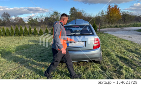 A Caucasian male mechanic in a gray and orange jacket opening the trunk of a silver car parked on a grassy roadside under a clear blue sky A Caucasian male mechanic in a gray and orange jacket opening the trunk of a silver car parked on a grassy roadside under a clear blue sky 134021729