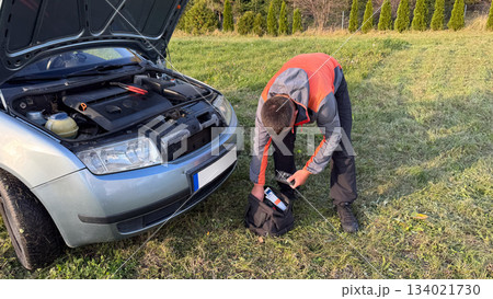 A Caucasian male mechanic in an orange and gray jacket retrieving a glow plug tester from a black tool bag next to a silver car with the hood open on a grassy roadside 134021730