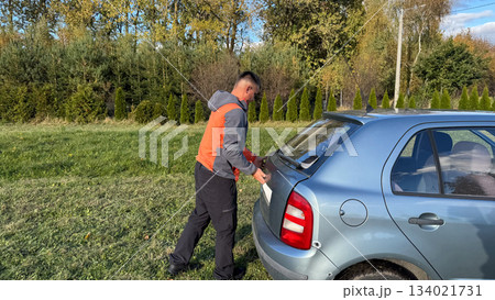 A Caucasian male mechanic in an orange and gray jacket closing the trunk of a silver car while standing on a grassy roadside in sunny weather A Caucasian male mechanic in an orange and gray jacket closing the trunk of a silver car while standing on a grassy roadside in sunny weather 134021731