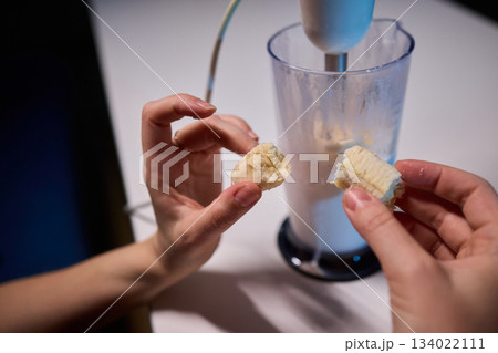 Hands prepare creamy banana smoothie, Electric blender mixes banana and milk for smoothie, Closeup of hands combining banana and milk to create refreshing breakfast drink 134022111
