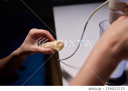 Hands prepare creamy banana smoothie, Electric blender mixes banana and milk for smoothie, Closeup of hands combining banana and milk to create refreshing breakfast drink 134022113