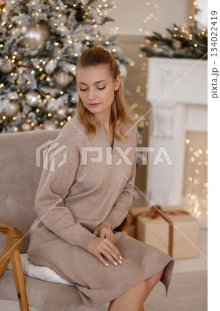 Caucasian woman sitting on chair in cozy Christmas room with tree and white fireplace 134022419