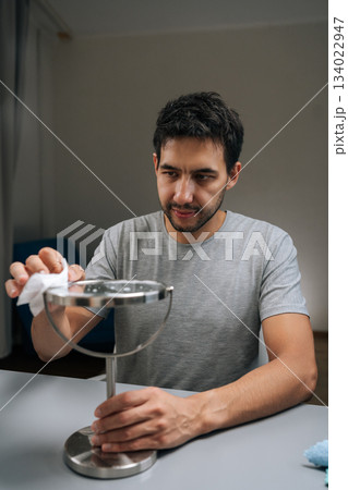 Vertical portrait of man using blue microfiber cloth to carefully clean round tabletop mirror, performing household chore with focus on personal hygiene and maintaining clean environment. Vertical portrait of man using blue microfiber cloth to carefully clean round tabletop mirror, performing household chore with focus on personal hygiene and maintaining clean environment. 134022947