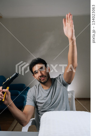 Vertical shot of curious man feeling bored with everyday chores, playfully engaging with steam cleaner, shooting vapor and raising hand, distracting himself from household cleaning routine. 134023063